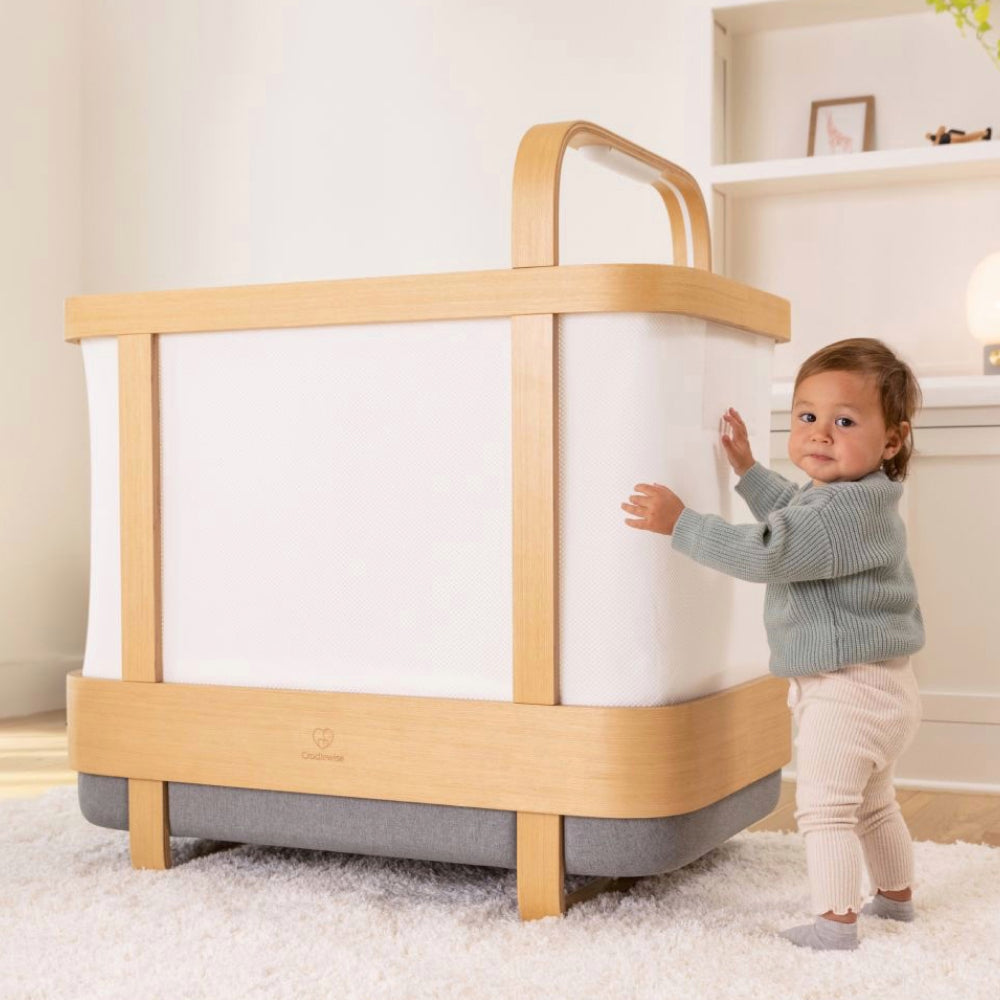 Toddler in a gray sweater standing beside a Cradlewise smart crib in crib mode, with one hand resting on the crib’s side panel.