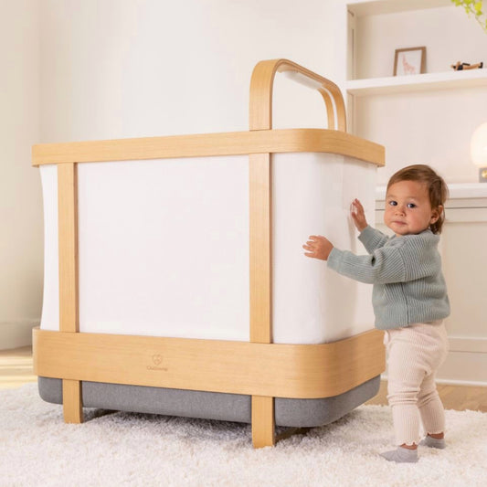 Toddler in a gray sweater standing beside a Cradlewise smart crib in crib mode, with one hand resting on the crib’s side panel.