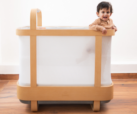Smiling baby standing inside a Cradlewise smart crib in crib mode, holding the light wooden top rail while looking forward, with white mesh panels fitted around the crib sides.