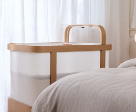 Cradlewise smart crib in bassinet mode with white hexagonal-pattern mesh panels attached to the wooden frame, positioned beside a bed in a bright bedroom with natural light filtering through sheer white curtains.