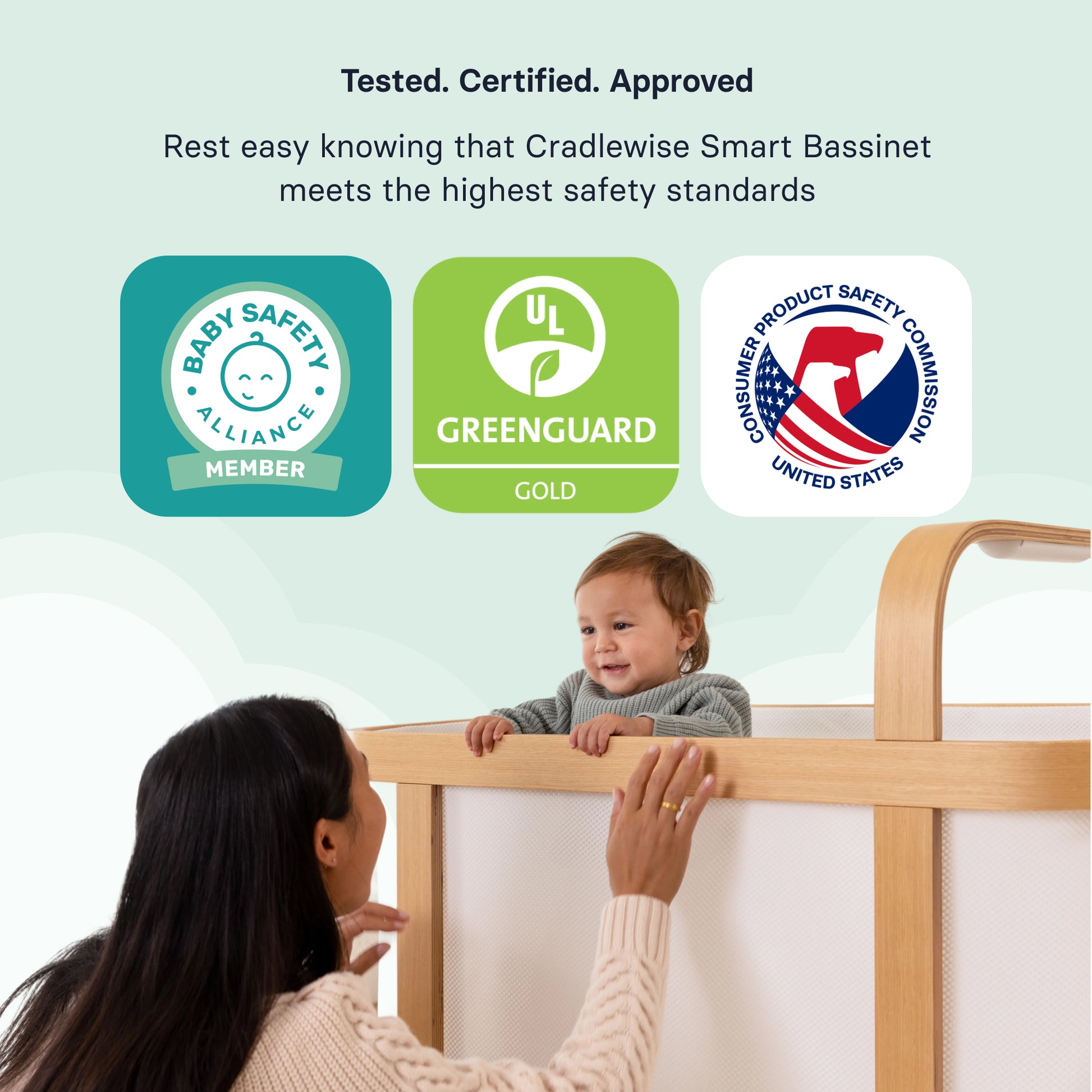 Parent watching a baby stand in the Cradlewise crib alongside Baby Safety Alliance, GREENGUARD Gold, and CPSC safety badges highlighting the crib’s safety certifications.