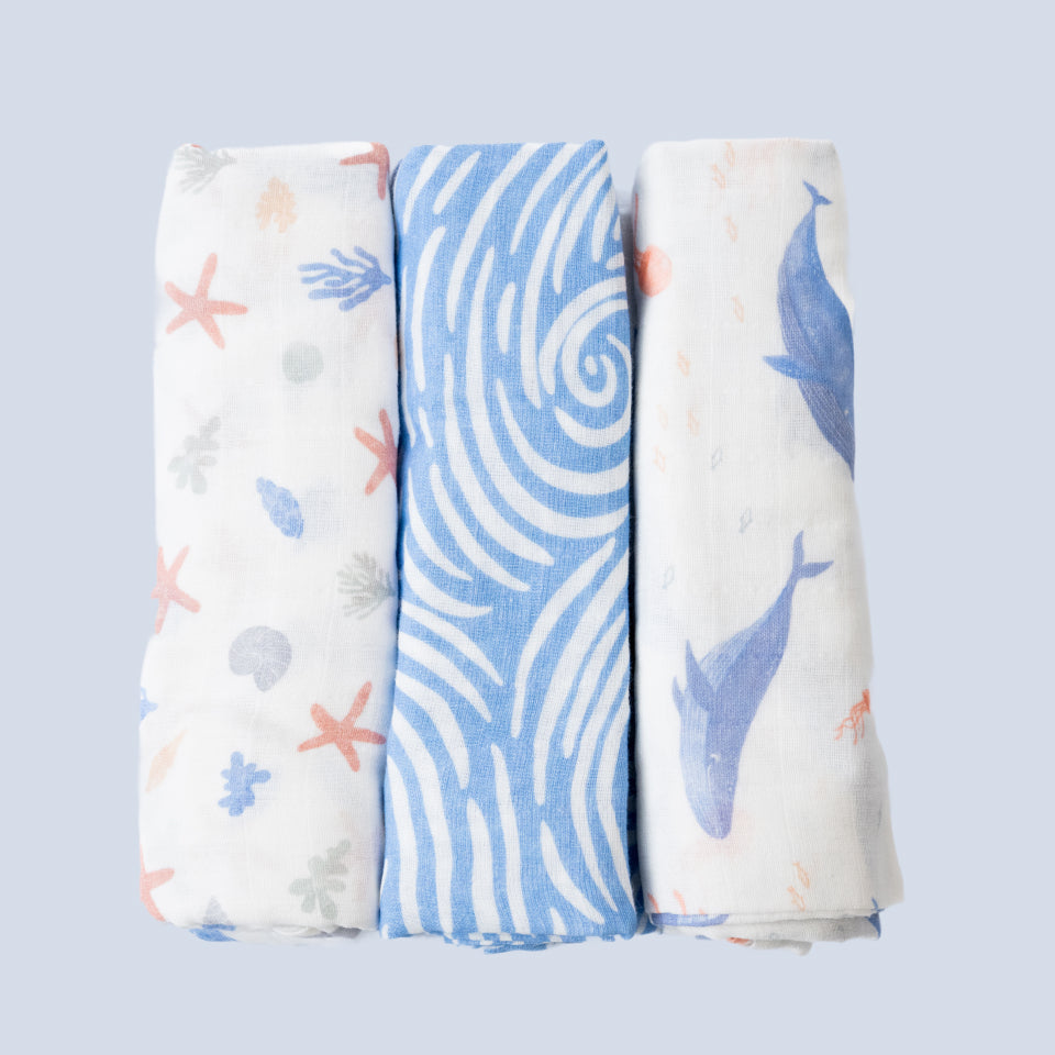 Three rolled Cradlewise Muslin Swaddles in Underwater Life, featuring a white swaddle with pastel starfish, seashells and coral motifs, a blue swaddle with white swirling wave patterns, and a white swaddle with blue whale motifs and small sea-inspired details.