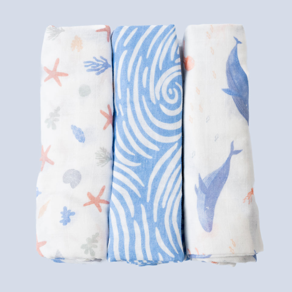 Three rolled Cradlewise Muslin Swaddles in Underwater Life, featuring a white swaddle with pastel starfish, seashells and coral motifs, a blue swaddle with white swirling wave patterns, and a white swaddle with blue whale motifs and small sea-inspired details.